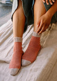 Person wearing pink socks with gray toes, sitting on a bed, next to a pink background with 'project 12 RACHEL Simple Socks' text.