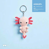 A knitted axolotl keychain shown next to a knitting pattern page. The keychain is light pink with red pectoral fins and a silver metal key ring.