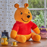 A crocheted Winnie the Pooh plush toy is displayed on a tabletop. The plush is orange with a red shirt and black nose, with small accessories including a honey pot and teacup.