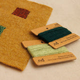 A close-up of a mustard yellow woven fabric swatch with three small woven color squares, next to two spools of thread labeled .nd in dark green and pale yellow on a light beige background.
