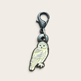 An owl-shaped metal charm with a lobster clasp, featuring a pale yellow enamel fill and dark metal outlines, shown on a light beige background.