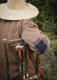 Person wearing a brown cardigan and straw hat sitting on a wooden chair with a close-up of the patch on the cardigan's sleeve.